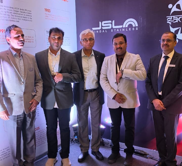 Five men in formal business attire posing at a JSW Stainless exhibition booth with Journey so far display in the background